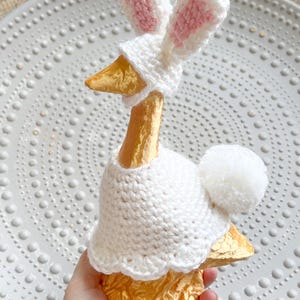 May include: A gold goose figurine wearing a white crocheted bunny costume with pink ears and a white pom-pom tail.
