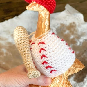 May include: A crocheted goose figurine wearing a red baseball cap and a white baseball jersey with red stitching. The goose is holding a crocheted baseball bat.
