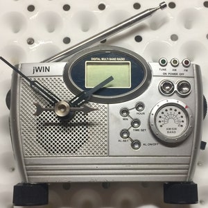 May include: A silver and black digital multi-band radio with a clock face and a telescopic antenna. The radio has a digital display, a dial for selecting the band, and buttons for tuning, setting the time, and turning the radio on and off. The radio also has a speaker grille on the front.