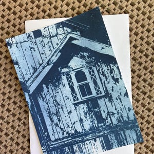 Może przedstawiać: Prostokątna kartka okolicznościowa z kontrastowym, monochromatycznym niebiesko-białym nadrukiem przedstawiającym fasadę zniszczonego drewnianego budynku. Obraz ukazuje linię dachu dwuspadowego, pionowe panele drewniane oraz centralne okno.