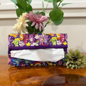 May include: Purple tissue box cover featuring a colourful print of cartoon bears. The bears are wearing different colours and have different expressions. The tissue box cover is made of fabric and has a purple trim.