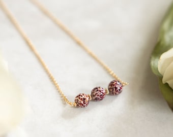 Op de afbeelding: Een gouden ketting met drie ronde, bordeauxrode en wit gevlekte kralen.