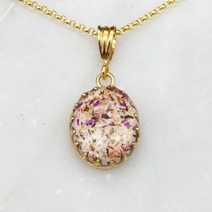 May include: A gold chain necklace with a pendant featuring a clear oval-shaped resin filled with dried flowers and a gold filigree border.