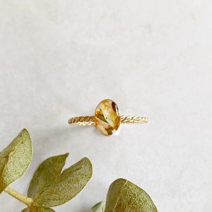 以下が含まれることがあります： ねじれたバンドと、押し花が入った楕円形のレジンストーンが付いたゴールドリング。花は白と黄色です。