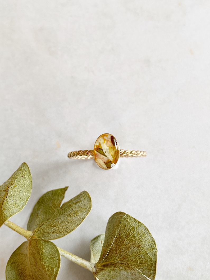 以下が含まれることがあります： ねじれたバンドと、押し花が入った楕円形のレジンストーンが付いたゴールドリング。花は白と黄色です。