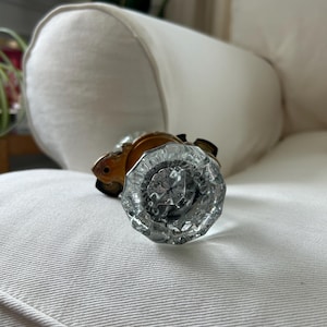 May include: A clear, faceted glass doorknob with a brass base. The knob is round with a cut-glass design, reflecting light. The hardware is likely for a door or cabinet, adding a vintage touch to home decor.