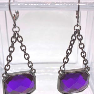 May include: A pair of dangle earrings featuring faceted, rectangular purple gemstones set in dark metal frames. The gemstones are suspended by a chain of small, dark metal links and attached to a lever-back clasp.