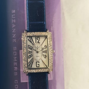 Puede incluir: Un reloj de cuarzo rectangular con una correa azul marino. La esfera es blanca con números romanos negros y un pequeño dial de segundos. El bisel está incrustado con pequeños cristales transparentes. El reloj es de la colección Suzanne Somers.