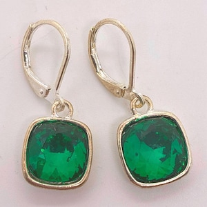May include: A pair of silver-toned leverback earrings. Each earring features a square, emerald-green gemstone set in a gold-toned frame. The gemstones have a faceted cut, reflecting light.