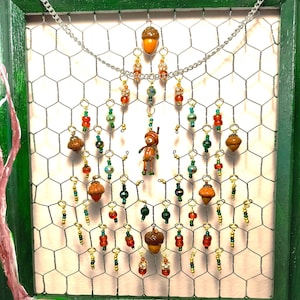 Può includere: Un'applique da parete decorativa con una cornice verde, rete metallica e ornamenti a tema ghianda. Gli ornamenti includono ghiande in miniatura, dettagli di perline rosse, verdi e dorate e una piccola statuina. Una catena d'argento è appesa in alto.