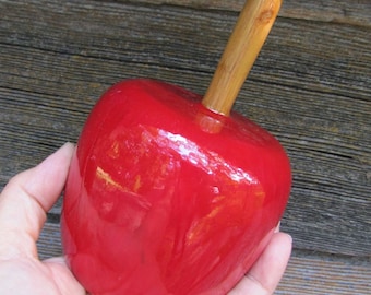 Könnte beinhalten: Ein roter, glänzender Keramikapfel mit einem Holzstiel. Der Apfel wird in einer Hand gehalten.