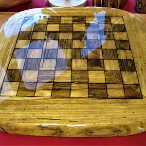 May include: A wooden chessboard with a clear epoxy resin finish. The board is made of light and dark wood, creating a checkered pattern. The board is oval shaped and has a natural wood edge.