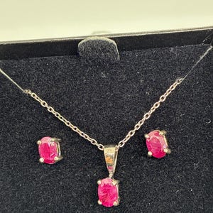 May include: A jewelry set featuring a silver necklace with a pendant and matching stud earrings. Each piece showcases a vibrant, oval-cut ruby gemstone. The set is presented on a black velvet surface within a black box.