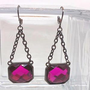 May include: A pair of dangle earrings featuring faceted, rectangular magenta-colored gems set in dark metal frames. The gems are suspended from a chain and hook closure.