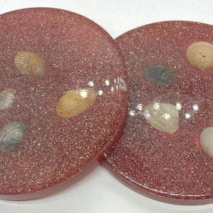 Puede incluir: Dos posavasos redondos de resina roja con conchas incrustadas. Los posavasos tienen un acabado brillante y presentan varias conchas pequeñas en tonos blancos, beige y grises. Estos posavasos decorativos están diseñados para proteger las superficies de las marcas de bebidas.
