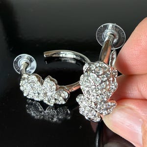 May include: A pair of silver hoop earrings with a floral design. Each earring is adorned with clear crystals. The earrings are held by a hand against a dark background.