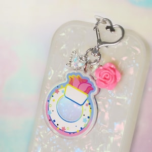 May include: A white phone case with a colorful charm attached. The charm features a pink flower, a clear round charm with a white and blue design, and a silver heart charm. The charm is attached to the phone case with a silver chain.