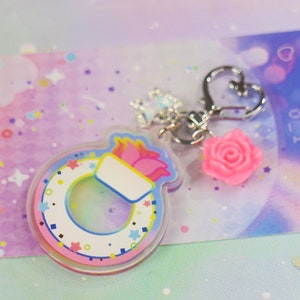 May include: A colorful paintbrush charm with a pink rose and a silver heart charm attached to a silver chain. The charm is made of clear acrylic and has a rainbow design.