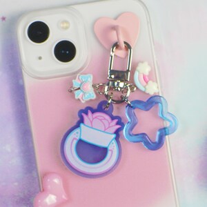 May include: A clear smartphone case with a pink gradient, featuring a silver metal keychain attachment. The keychain includes a pink heart, a small blue and pink bow, a tiny rainbow, a blue star-shaped charm, and a purple ring-shaped charm.