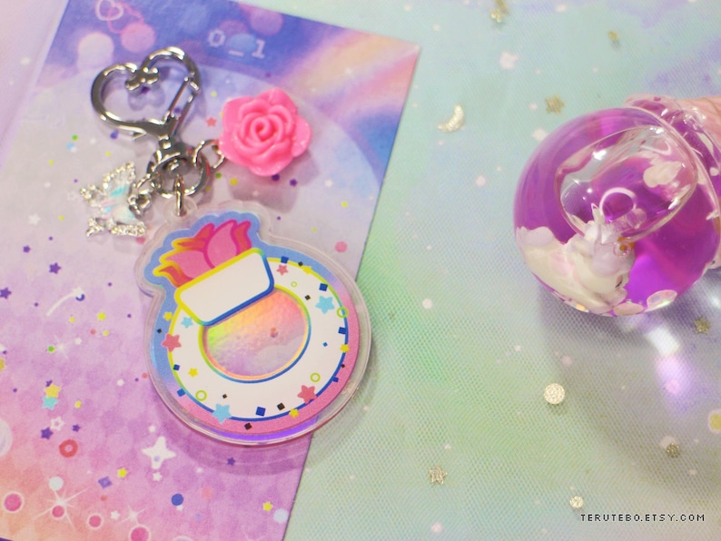 May include: A pink and white charm with a rainbow swirl inside, attached to a silver key chain with a pink flower charm. The charm is shaped like a ring with a pink flower on top. The charm is on a pink and purple background with white stars.