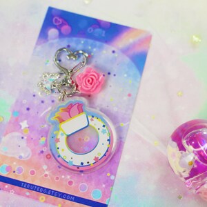 May include: A pink and white keychain charm with a flower and a life preserver design. The charm is attached to a silver key ring with a heart-shaped charm. The background is a rainbow gradient with glitter.
