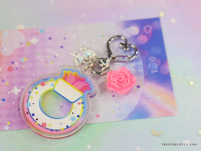 May include: A colorful paintbrush charm with a pink rose and a silver heart charm attached to a silver chain. The charm is made of clear acrylic and has a rainbow design.