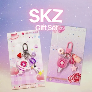 Puede incluir: Dos llaveros en tarjetas de presentación con el texto SKZ Gift Set. El llavero izquierdo tiene un dije de barra de luz blanca, un corazón rojo, una cuenta morada y un astronauta. El llavero derecho tiene una barra de luz rosa con forma de gato.