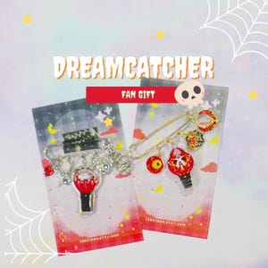 May include: Two silver-toned safety pins featuring enamel charms on display cards. The left pin has a red and white lightstick charm and a skull. The right pin features a red eye, a red and white lightstick, a spiderweb, and a red crescent moon.
