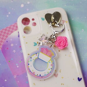 May include: A white phone case with a pink and blue charm attached. The charm features a ring with a pink flower and a white background. The phone case has a glittery finish and a heart-shaped cutout.