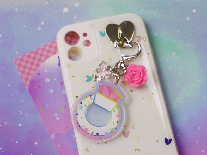 May include: A white phone case with a pink and blue charm attached. The charm features a ring with a pink flower and a white background. The phone case has a glittery finish and a heart-shaped cutout.