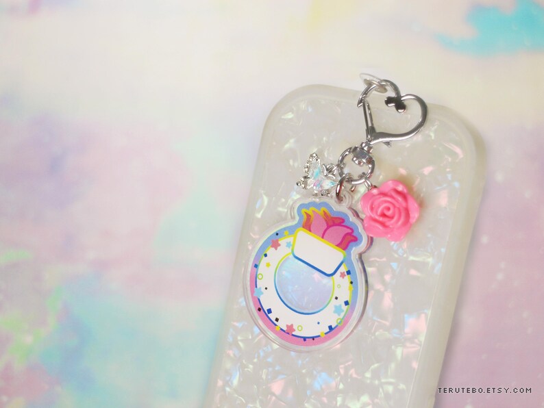 May include: A white phone case with a colorful charm attached. The charm features a pink flower, a clear round charm with a white and blue design, and a silver heart charm. The charm is attached to the phone case with a silver chain.