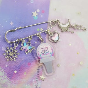 Puede incluir: Un broche de seguridad plateado con abalorios, incluyendo un copo de nieve, una mariposa, un corazón, una luna creciente y un abalorio transparente con un logotipo rosa y azul que dice "æ".