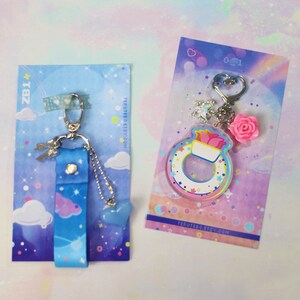 May include: Two keychains with a pastel rainbow and cloud theme. One keychain features a blue and white fabric strap with a silver charm. The other keychain features a clear acrylic charm with a rainbow and white design. Both keychains have a silver ring.