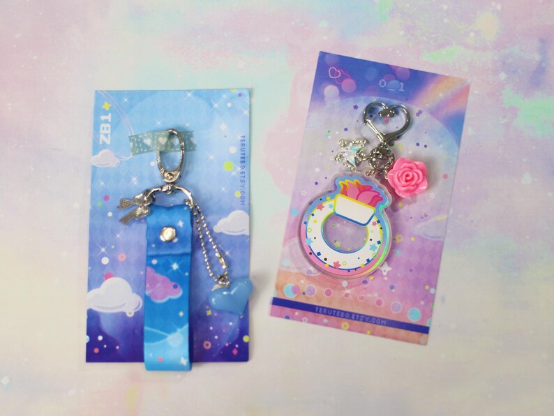 May include: Two keychains with a pastel rainbow and cloud theme. One keychain features a blue and white fabric strap with a silver charm. The other keychain features a clear acrylic charm with a rainbow and white design. Both keychains have a silver ring.