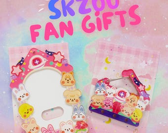 May include: Two acrylic items featuring SKZOO character illustrations on pink gingham backing cards. The left item is a vertical frame with a central cutout, and the right is a smaller horizontal frame. Text at the top reads SKZOO FAN GIFTS.