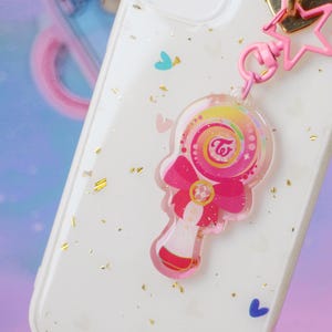 Op de afbeelding: Een witte telefoonhoes met gouden glitter en een roze en regenboogkleurige hanger. De hanger is een lolly met een roze strik en de tekst "TWICE" in een witte cirkel.