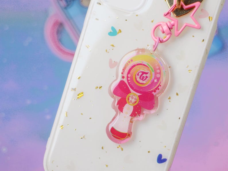 Op de afbeelding: Een witte telefoonhoes met gouden glitter en een roze en regenboogkleurige hanger. De hanger is een lolly met een roze strik en de tekst "TWICE" in een witte cirkel.