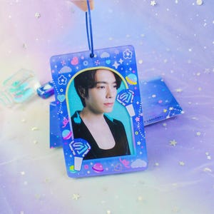 Puede incluir: Un colgante decorativo rectangular azul con un retrato de una persona. El colgante está decorado con corazones, estrellas y planetas de dibujos animados. Tiene una cuerda azul para colgar y dos diseños de micrófono estilizados.