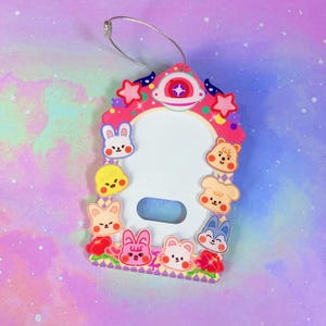 May include: A pink and white ID badge holder with a clear window. The holder is shaped like a house and decorated with cartoon animal faces, stars, and a spaceship. A silver wire loop is attached at the top.