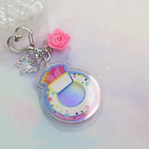 May include: A colorful keychain with a pink flower charm, a silver heart-shaped ring, and a clear acrylic charm with a rainbow iridescent background and a white inner circle with a pink and blue flower design. The charm has a white space for personalization.