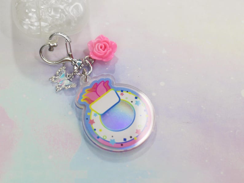 May include: A colorful keychain with a pink flower charm, a silver heart-shaped ring, and a clear acrylic charm with a rainbow iridescent background and a white inner circle with a pink and blue flower design. The charm has a white space for personalization.