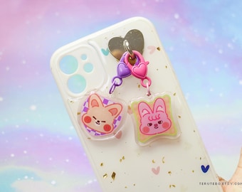 May include: A clear phone case with gold flecks and a heart-shaped metal charm. Attached are two acrylic charms: one round with a fox design and one square with a bunny design. The charms are pink, purple, and yellow.
