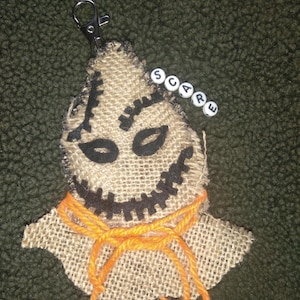 May include: A burlap keychain with a smiling face design, featuring black stitched details and an orange yarn tie. The word "scare" is spelled out in white letter beads. It has a silver clasp.