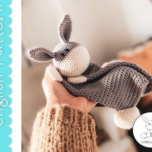 Puede incluir: Un juguete de burro de crochet gris y blanco con una manta adjunta. El juguete está siendo sostenido por una mano que lleva un suéter marrón.