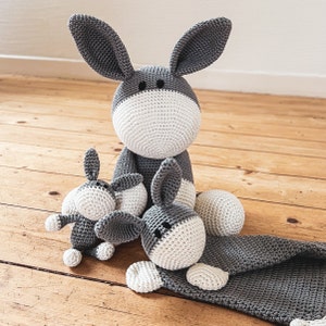 Può includere: Tre peluche di asino in crochet grigio e bianco. L'asino più grande è seduto con le gambe distese. I due asini più piccoli sono seduti davanti al più grande.