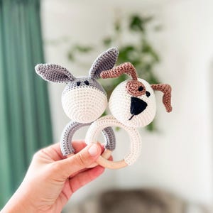 Könnte beinhalten: Ein gehäkeltes Babyrassel mit einem grauen Hasen und einem braunen und weißen Hund, die an einem Holzring befestigt sind.