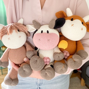 May include: Three crocheted cow plushies in different colors. The brown cow has a yellow flower on its head. The pink cow has a white face and black spots. The white cow has a brown face and brown spots.
