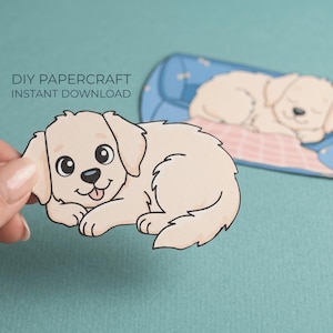 May include: A papercraft illustration of a light beige puppy with black eyes and a pink tongue. The puppy is lying down, and a second puppy is sleeping on a blue dog bed. Text reads "DIY Papercraft Instant Download."
