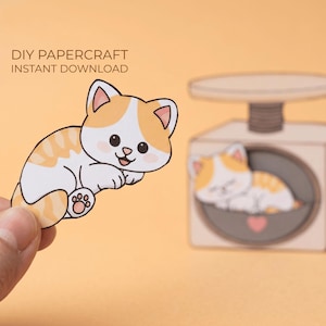 Könnte beinhalten: Ein DIY-Papierbastelprojekt mit einer Cartoon-Katze mit orangefarbenen und weißen Markierungen. Die Katze befindet sich neben einer Papierwaage mit einer schlafenden Katze im Inneren. Der Text "DIY PAPER CRAFT INSTANT DOWNLOAD" steht oben.