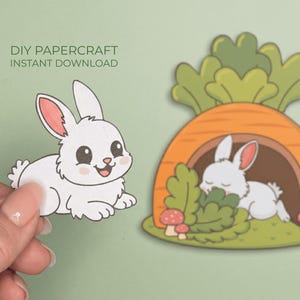 Könnte beinhalten: Ein weißes Papierkaninchen mit rosa Innenohren und einem lächelnden Gesicht wird von einer Hand gehalten. Daneben eine Cartoon-Illustration eines Kaninchens, das in einem Karottenhaus schläft. Text: DIY PAPERCRAFT INSTANT DOWNLOAD.